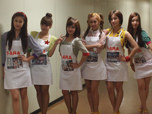 “Ｔ－ＡＲＡエプロン”を身に付けてポーズを取っているガールズグループのＴ－ＡＲＡ（ティアラ、写真＝コアコンテンツメディア提供）。