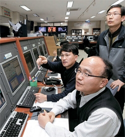 京畿道竜仁市のＫＴ衛星管制センターで、イ・ジェヨル管制運用チーム長（前）と職員がムグンファ５号から送られてきた信号を分析している。 職員は英単語と数字が組み合わされた信号を読み取り、軌道修正など人工衛星に必要な措置を取る。