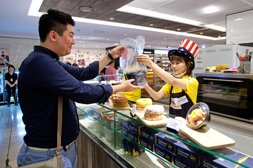 江南（カンナム）エリアを代表するデパート、現代百貨店貿易センター店の地下食品売り場。ポップなユニフォームのスタッフから、男性が何かを受け取っています。カウンターには丸いお菓子のようなものが見えますが…。 