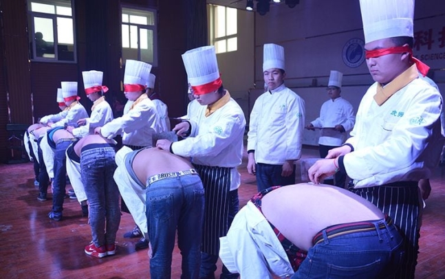 中国湖北省にある虎振技術学校料理学科の生徒が目隠しをしたままクラスメイトの背中の上で包丁を使っている様子。［写真　虎振学校公式サイト］