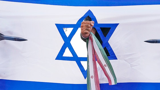 ２４日（現地時間）、イランの首都テヘランの市民がイスラエルの自国領土攻撃に抗議するため、イランの国旗を手首に結んだ拳をイスラエルの国旗から突き出すパフォーマンスをしている。　［ＡＰ＝聯合ニュース］