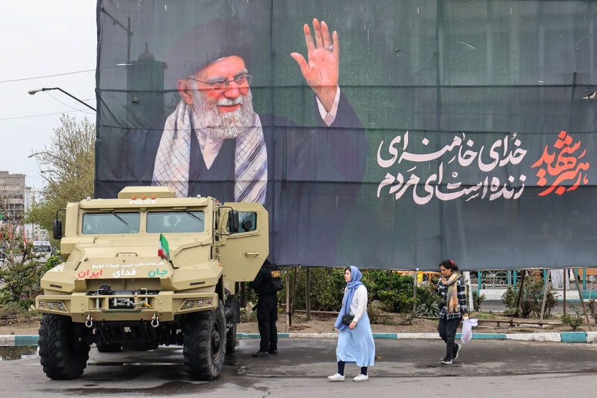 イランの首都テヘランで3月31日、ハメネイ師を追悼するバナーの下でイラン保安軍の車両のそばを女性らが歩いている。 ［AFP＝聯合ニュース］