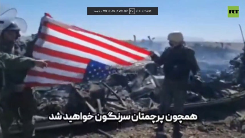 5日（現地時間）、イラン国営放送IRIBは、航空機墜落現場で弾薬箱を回収するイラン人が米国旗を逆さに掲げる場面を放送した。下には「自国の国旗のように転覆するだろう」との字幕が表示された。［写真　RT キャプチャー］