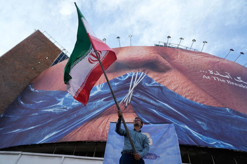 6日、イラン・テヘランのヴァリアスル広場で、ある男性がイラン国旗を振っている。男性の後ろには、ドナルド・トランプ米大統領の口をホルムズ海峡を象徴する青い布で塞いで糸で縫い合わせたような絵の看板が掲げられている。AP＝聯合ニュース