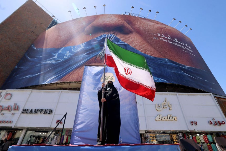 10日（現地時間）、イラン・テヘランのバリアスル広場で、女性がイランの国旗を手にしている。女性の後ろにはトランプ米大統領の口をホルムズ海峡を象徴する青い布で塞ぎ、糸で縫い合わせたような絵の看板が掲げられている。［AFP＝聯合ニュース］