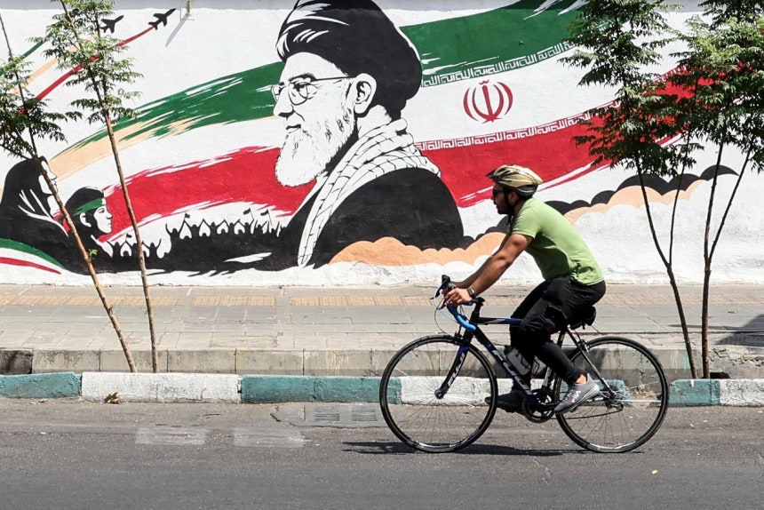 24日、イラン・テヘランで、故アヤトラ・アリ・ハメネイ前最高指導者とイラン国旗が描かれた壁画の前を、自転車に乗った市民が通り過ぎている。AFP＝聯合ニュース